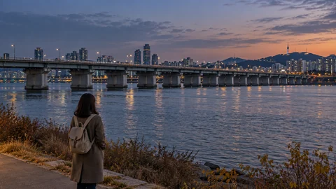 A traveler in an autumn coat looking across a quiet river toward a low bridge at dusk