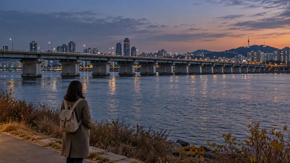 A traveler in an autumn coat looking across a quiet river toward a low bridge at dusk