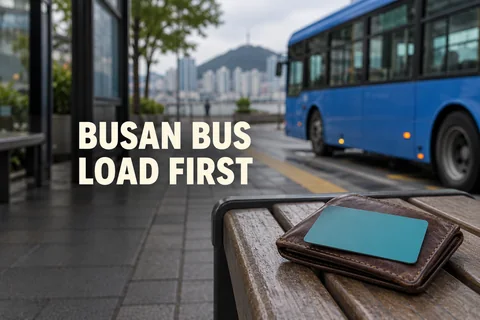 A plain transit card and wallet at an imagined bus stop, with an out-of-focus bus and the editorial reminder BUSAN BUS: LOAD FIRST.