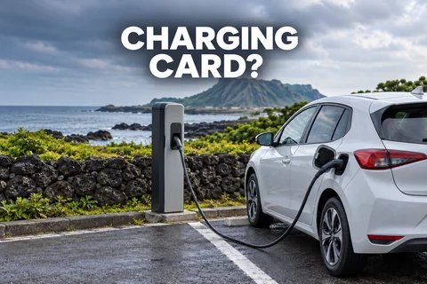 An electric hatchback connected to a charging pedestal beside a coastal stone wall, with the editorial text CHARGING CARD?