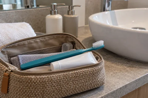 A toothbrush and small toothpaste tube in a travel pouch beside a hotel washbasin and refillable pump bottles