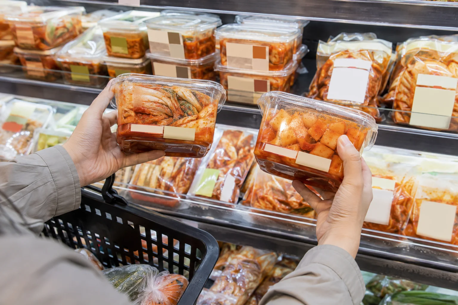 A shopper comparing unbranded napa cabbage and cubed radish kimchi containers in a refrigerated aisle
